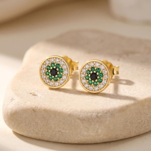 May include: A pair of round gold stud earrings featuring a central black stone surrounded by a ring of small green stones and an outer border of clear white stones. The circular design creates a layered, multi-colored effect on each earring.