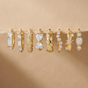 Könnte beinhalten: Eine Auswahl an goldenen Creolen mit verschiedenen Designs, darunter klare Edelsteine im Baguette- und Rundschliff, eine geflochtene Textur, eine Herzform und ein Sternmuster. Die Ohrringe werden vor einem neutralen Hintergrund präsentiert.
