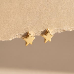 May include: A pair of gold-colored star-shaped stud earrings. The earrings are small and have a polished finish. They are displayed on a textured, light brown surface.