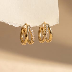May include: A pair of gold hoop earrings, each featuring a double-layered design. One layer is a smooth, polished gold band, while the second layer is a thinner gold band encrusted with a row of small, clear, sparkling stones along the front.