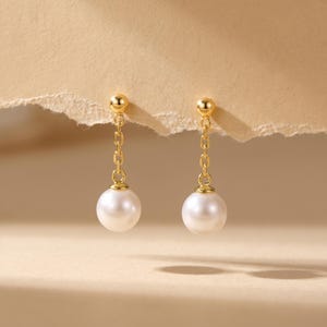 May include: A pair of gold-coloured drop earrings. Each earring has a small gold ball at the top, connected by a delicate chain to a large, round, white pearl. The earrings are displayed against a neutral background.