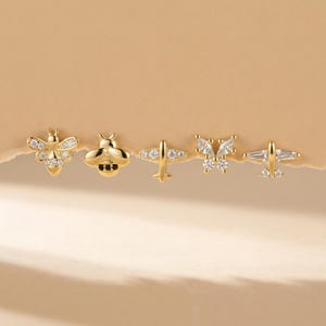 Puede incluir: Colección de cinco pendientes de oro. Los pendientes presentan varios diseños, incluyendo una abeja, una mariposa y dos diseños de avión. Los pendientes están adornados con pequeñas gemas transparentes, añadiendo brillo a la joyería.
