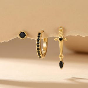 May include: A set of three gold-colored earrings. One is a stud with a black stone, another is a hoop with black stones, and the third is a dagger-shaped earring with black stones.