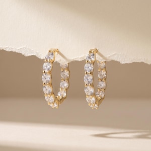 May include: A pair of gold hoop earrings featuring a continuous row of round, clear, faceted stones set along the front-facing edge of each circular band. The earrings are constructed with a polished gold-tone metal finish and clear stones.