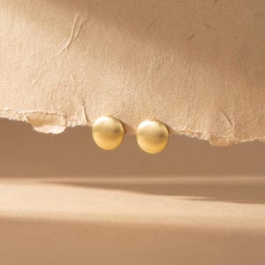 May include: A pair of gold-coloured, circular stud earrings. The earrings have a brushed metal finish and are presented on a textured, light brown surface. The background is a soft, neutral tone.