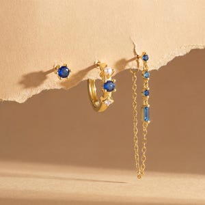 Könnte beinhalten: Eine Auswahl an goldenen Ohrringen mit blauen Edelsteinen. Das Set umfasst einen Ohrstecker, einen Creolen-Ohrring mit einer Perle und einen Hängeohrring mit Kette. Die Ohrringe werden vor einem neutralen Hintergrund präsentiert.