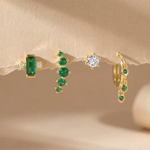 May include: Four gold earrings with emerald and diamond accents. One earring features a rectangular emerald, while another has a cluster of emeralds. A third earring has a single diamond, and the last is a hoop with emerald drops.