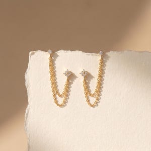 May include: A pair of gold earrings featuring a small, four-pointed star-shaped stud set with a clear stone. Each stud is connected to a delicate double chain that loops back to a second, smaller four-pointed star-shaped stud with a clear stone.
