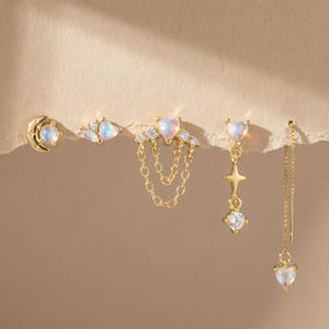 May include: A collection of gold-toned earrings featuring various designs. Styles include a crescent moon with a gemstone, a cluster of gemstones, heart-shaped studs with chain accents, a star and diamond drop, and a delicate chain with a heart charm.