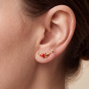 May include: Gold-colored koi fish earrings with red enamel detailing. The earrings are shaped like swimming koi fish, with the red enamel highlighting the fish's markings. The earrings are shown on an ear.