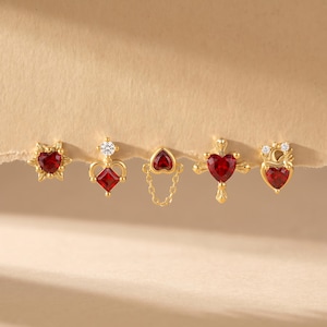 Könnte beinhalten: Eine Sammlung von fünf goldfarbenen Ohrringen mit roten herzförmigen Edelsteinen. Die Ohrringe weisen verschiedene Designs auf, darunter ein Sonnenstrahl, eine Krone, ein verbundenes Herz, ein Kreuz und ein Herz mit einer Krone. Jeder Ohrring hat eine einzigartige Form.