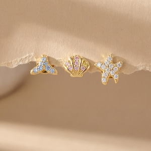 May include: Three gold-colored stud earrings in a nautical theme. The earrings are shaped like a whale tail, seashell, and starfish. The whale tail is blue, the seashell is pink and yellow, and the starfish is white. The earrings are set against a neutral background.