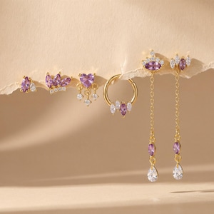 Op de afbeelding: Een collectie goudkleurige oorbellen met paarse en heldere edelstenen. De oorbellen omvatten studs, hoops en hangende stijlen met verschillende vormen, waaronder harten, kronen en druppels. De sieraden worden op een neutrale achtergrond gepresenteerd.