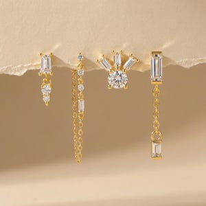 Puede incluir: Cuatro pendientes dorados con piedras de circonita cúbica transparentes y brillantes. Los diseños incluyen una piedra rectangular colgante con piedras más pequeñas, una cadena de piedras, un diseño de sol y una piedra rectangular colgante con una cadena.