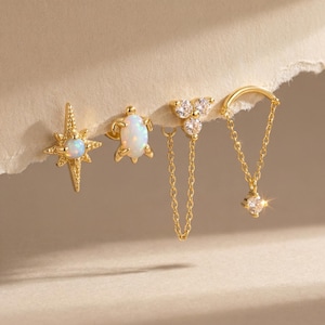 May include: A collection of gold-toned earrings featuring various designs. Included are a starburst stud with a central opal, a turtle stud with an opal shell, a cluster of three clear stones with a chain, and a chain earring with a dangling diamond-like stone.
