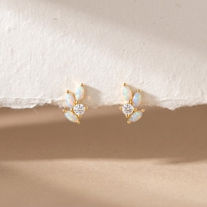 May include: A pair of gold-colored stud earrings. Each earring features a central, round, clear gemstone surrounded by three opal-colored, marquise-cut gemstones. The earrings are set against a neutral background.