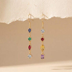 Op de afbeelding: Twee goudkleurige oorhangers, elk met een reeks kleurrijke edelstenen. De edelstenen zijn blauw, groen, rood, geel en paars in verschillende vormen. De oorbellen hangen aan een delicate gouden haak.