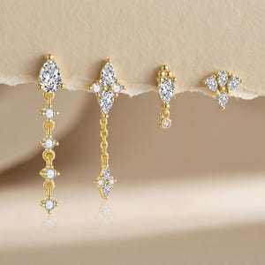 Puede incluir: Cuatro pares de pendientes dorados con piedras de circonita cúbica transparentes. Los diseños incluyen gotas colgantes, una forma de cruz con una cadena, una pequeña gota con un círculo colgante y un grupo de tres gotas.