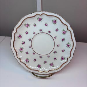 May include: A white porcelain saucer with a scalloped edge, adorned with small pink rose floral patterns and a gold trim. The center has a circular indentation. The saucer is displayed on a gold-colored metal stand.