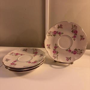 以下が含まれることがあります： バラの花柄が特徴のヴィンテージスタイルのソーサーのセット。ソーサーは白地にピンクのバラと金の縁取りが施されています。1つのソーサーは金色のスタンドに、3つのソーサーのスタックが左側にあります。