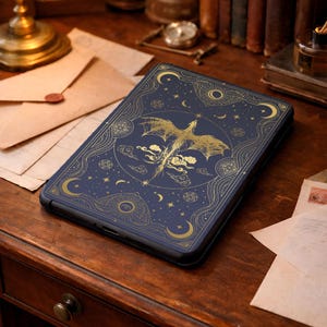 May include: A dark blue e-reader cover featuring a gold-colored line art illustration of a dragon in flight, surrounded by celestial motifs including crescent moons, stars, and clouds, with intricate geometric patterns framing the central design.