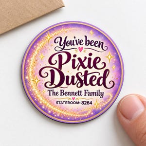 May include: A circular sticker with a purple and cream gradient background featuring gold glittery star accents and a swirling trail. The text reads You've been Pixie Dusted, The Bennett Family, and STATEROOM: 8264 in dark purple script and print.