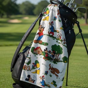May include: A black golf bag with golf clubs and a white towel featuring cartoon characters. The towel has a repeating pattern of Mickey Mouse, Minnie Mouse, Goofy, and Pluto on a golf course setting.