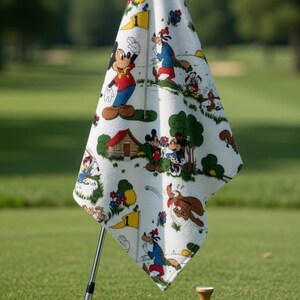 May include: A white golf towel featuring a colorful cartoon print of Mickey Mouse, Goofy, and other Disney characters. The towel is draped over a golf club, with a golf ball and tee in the foreground on a green golf course.