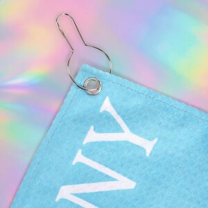 May include: A close-up of a light blue towel corner with white text and a silver metal hook. The towel has a stitched edge. The background is a colorful, iridescent pattern.