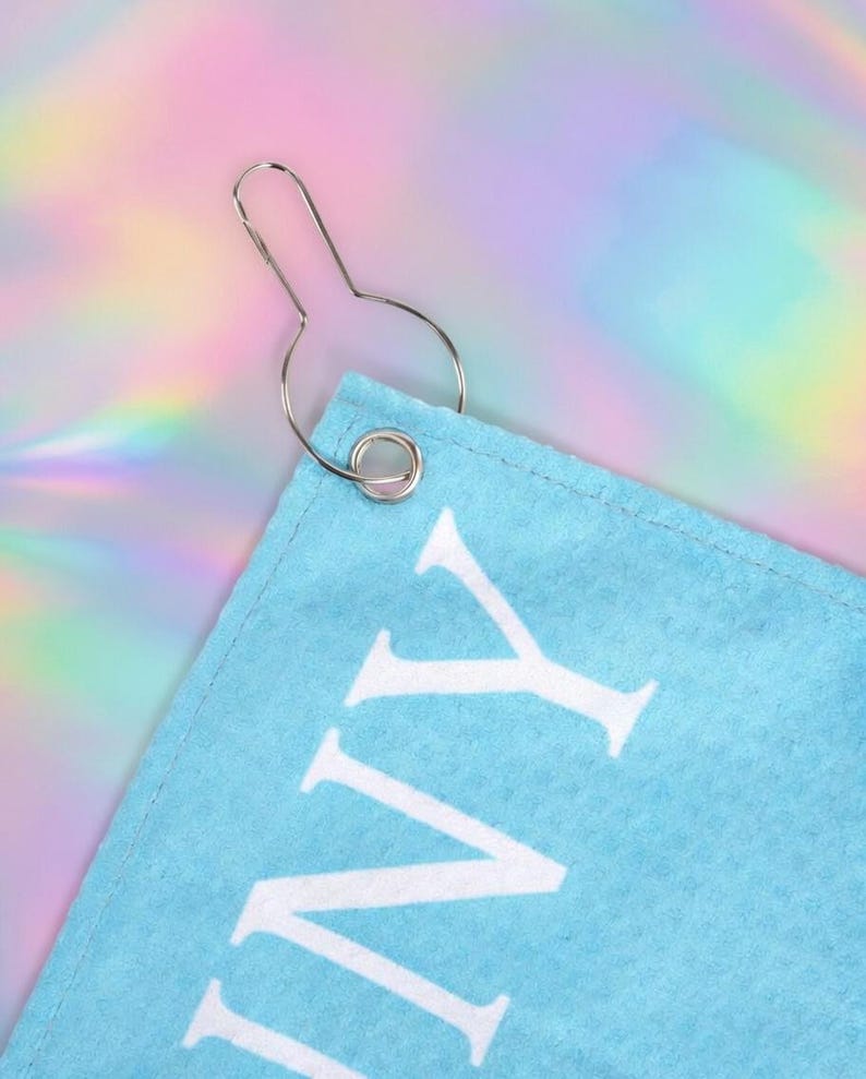 May include: A close-up of a light blue towel corner with white text and a silver metal hook. The towel has a stitched edge. The background is a colorful, iridescent pattern.