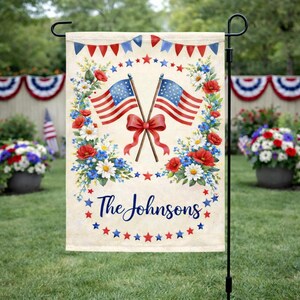 May include: A garden flag with two crossed American flags and a red bow, encircled by a floral wreath of red, white, and blue flowers. The text "The Johnsons" is below, with a star border. The flag is displayed on a black metal stand in a garden.