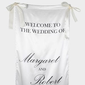 May include: A white satin fabric banner featuring black printed text that reads WELCOME TO THE WEDDING OF Margaret AND Robert. The banner is suspended by two white satin ribbons tied into bows at the top left and top right corners.