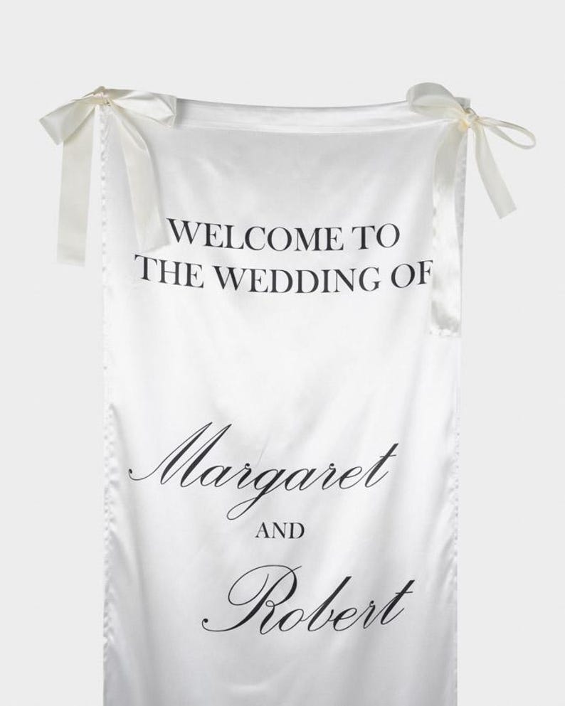 May include: A white satin fabric banner featuring black printed text that reads WELCOME TO THE WEDDING OF Margaret AND Robert. The banner is suspended by two white satin ribbons tied into bows at the top left and top right corners.