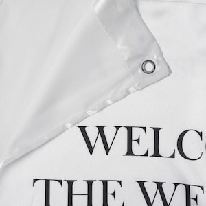 May include: A white fabric shower curtain featuring a reinforced header with a silver metal grommet. The fabric has a smooth, satin-like finish. Printed in bold black capital letters, the partial text reads WELC and THE WE.