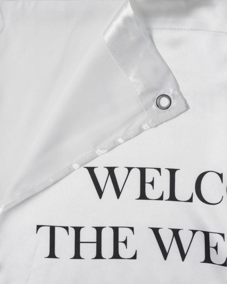 May include: A white fabric shower curtain featuring a reinforced header with a silver metal grommet. The fabric has a smooth, satin-like finish. Printed in bold black capital letters, the partial text reads WELC and THE WE.