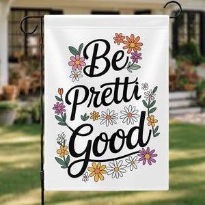May include: A white garden flag with the words "Be Pretti Good" in black script, surrounded by colorful flowers in orange, pink, yellow, and purple. The flag is displayed outdoors on a black metal stand.