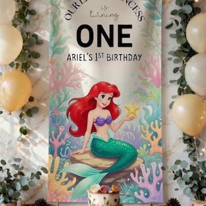 May include: A vertical rectangular banner featuring a cartoon mermaid with red hair and a green tail sitting on a rock, surrounded by colorful coral. The text reads: OUR LITTLE PRINCESS is turning ONE ARIEL'S 1ST BIRTHDAY. The background is light.