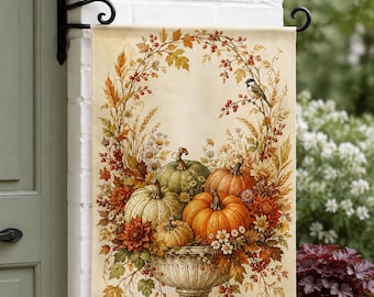 May include: A rectangular garden flag featuring a decorative urn filled with orange, green, and white pumpkins, surrounded by autumn leaves, berries, wheat, and flowers. A small bird is perched on a branch arching over the arrangement.