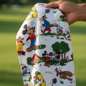 May include: White golf towel featuring a colorful cartoon print of Mickey Mouse, Minnie Mouse, Goofy, Donald Duck, and Pluto playing golf. The towel has a golf course scene with trees, flowers, and golf flags.