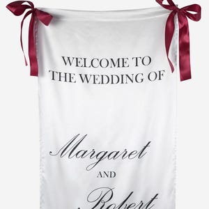 May include: A white fabric wedding welcome sign featuring the black printed text WELCOME TO THE WEDDING OF Margaret AND Robert. The rectangular banner is suspended by two dark burgundy satin ribbons tied into bows at the top corners.