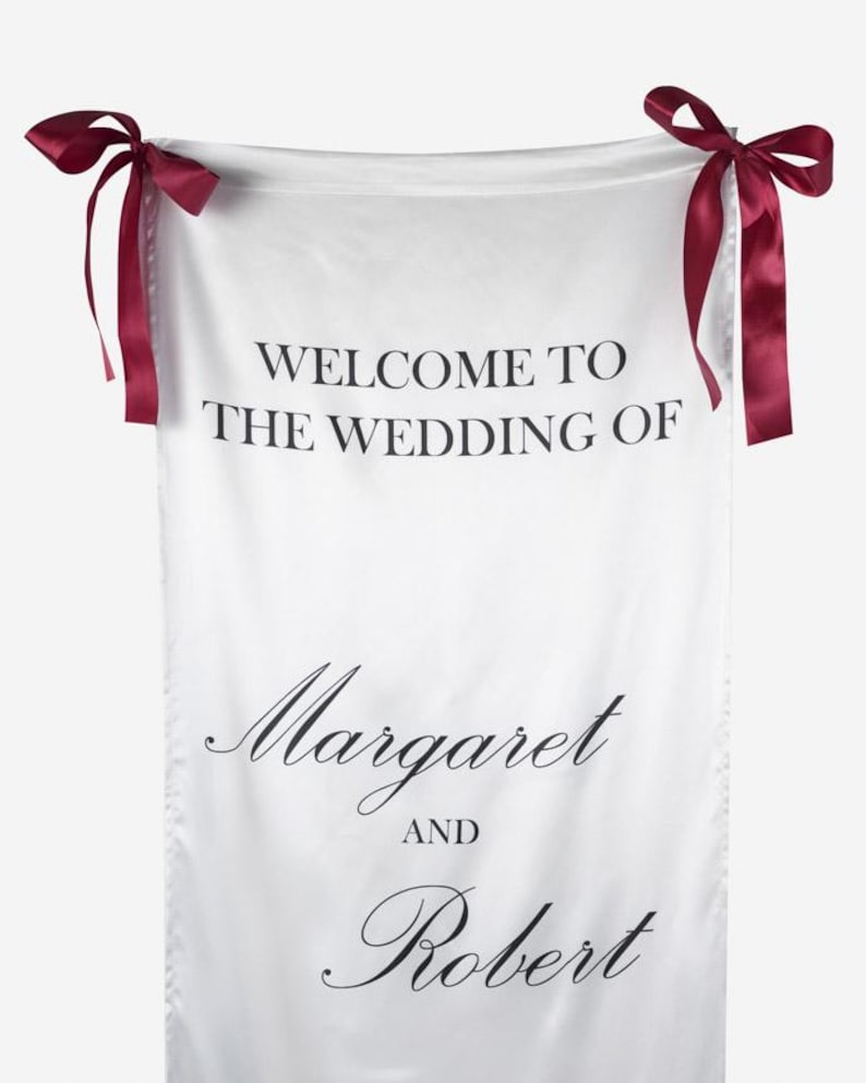 May include: A white fabric wedding welcome sign featuring the black printed text WELCOME TO THE WEDDING OF Margaret AND Robert. The rectangular banner is suspended by two dark burgundy satin ribbons tied into bows at the top corners.