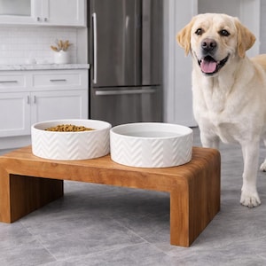 May include: A light-colored Labrador Retriever stands next to a wooden elevated pet feeder. The feeder features two round white ceramic bowls with a textured chevron pattern. One bowl contains brown dry kibble, and the other is filled with water.