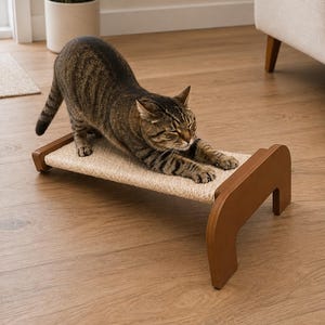 May include: A brown tabby cat stretches on a cat scratcher. The cat scratcher has a woven sisal surface and a wooden frame. The cat is brown and black with stripes. The cat scratcher is a light beige color.