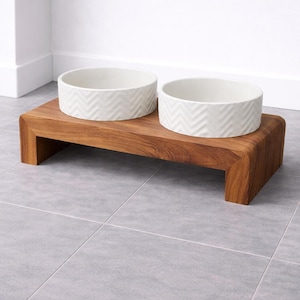 May include: A raised wooden stand with a natural brown wood grain finish, featuring two circular white ceramic bowls. Each bowl has a textured chevron pattern around the exterior. The stand has a rectangular shape with rounded edges and four legs.