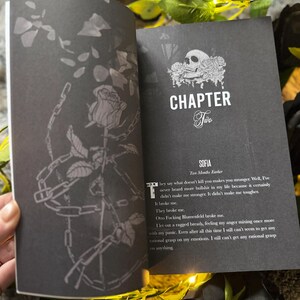 以下が含まれることがあります： 開いた本、濃い灰色のページ。左ページにはバラと鎖のイラスト。右ページにはテキストの上に「CHAPTER Two」と書かれています。テキストには「SOFIA」という名前と「Two Months Earlier」というフレーズが含まれています。
