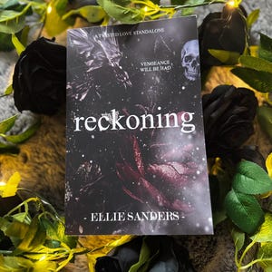 以下が含まれることがあります： Ellie Sandersの「reckoning」というタイトルの本。髑髏と花の要素が描かれた暗い表紙。黒いバラと緑の葉に囲まれ、フェアリーライトが飾られており、ゴシック様式を醸し出しています。