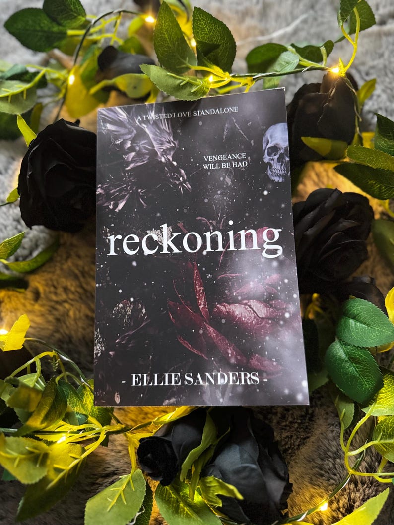 以下が含まれることがあります： Ellie Sandersの「reckoning」というタイトルの本。髑髏と花の要素が描かれた暗い表紙。黒いバラと緑の葉に囲まれ、フェアリーライトが飾られており、ゴシック様式を醸し出しています。
