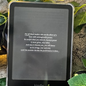 K&ouml;nnte beinhalten: Schwarzer Kindle E-Reader mit transparentem Schutzgeh&auml;use. Der Bildschirm zeigt Text &uuml;ber Macht und Entscheidungen. Das Wort "kindle" ist unten aufgedruckt. Gr&uuml;ne Bl&auml;tter und eine pelzige Oberfl&auml;che umgeben das Ger&auml;t.