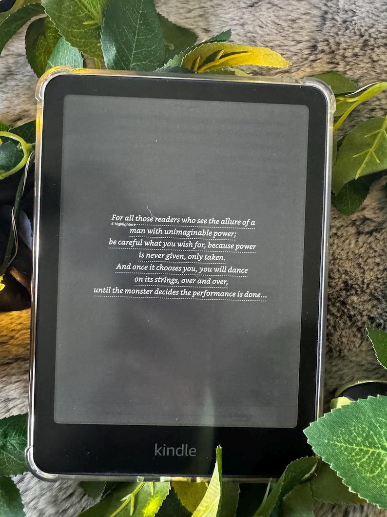 K&ouml;nnte beinhalten: Schwarzer Kindle E-Reader mit transparentem Schutzgeh&auml;use. Der Bildschirm zeigt Text &uuml;ber Macht und Entscheidungen. Das Wort "kindle" ist unten aufgedruckt. Gr&uuml;ne Bl&auml;tter und eine pelzige Oberfl&auml;che umgeben das Ger&auml;t.