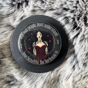May include: A circular black phone grip featuring an illustration of a person with long dark hair wearing a red corset. The text reads: Normal people dont understand how beautiful the darkness is. The design includes small red roses and vines.